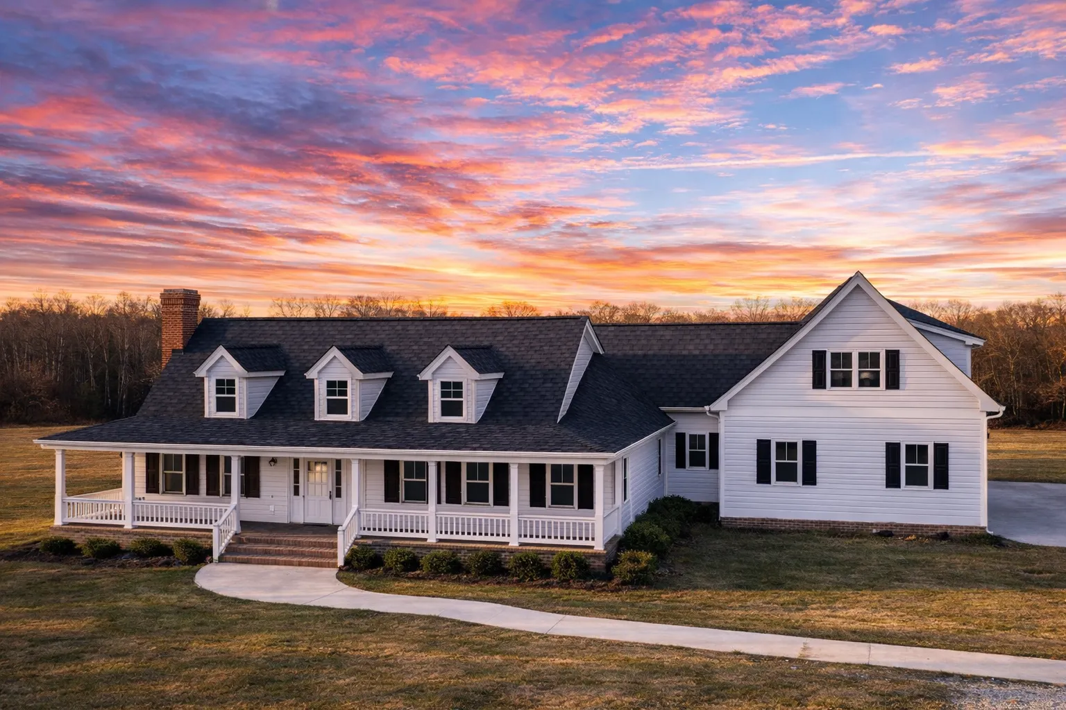 Front elevation of a Colonial Farmhouse style home with white lap siding, brick foundation, symmetrical windows, dormer accents, and a covered front porch