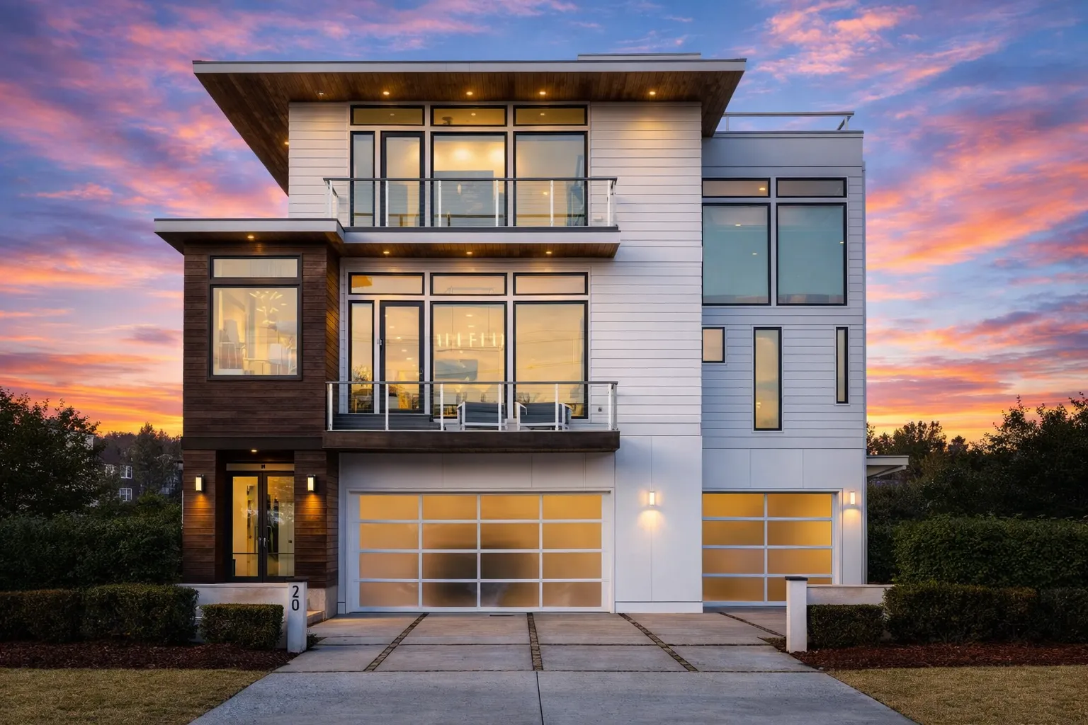 Front exterior view of a modern contemporary home with stucco and horizontal siding, glass balcony railings, flat rooflines, and warm interior lighting