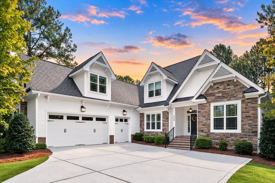 Front exterior of a Cape Cod cottage style home with horizontal siding, stone accents, steep gabled roof, and inviting red front door at sunset