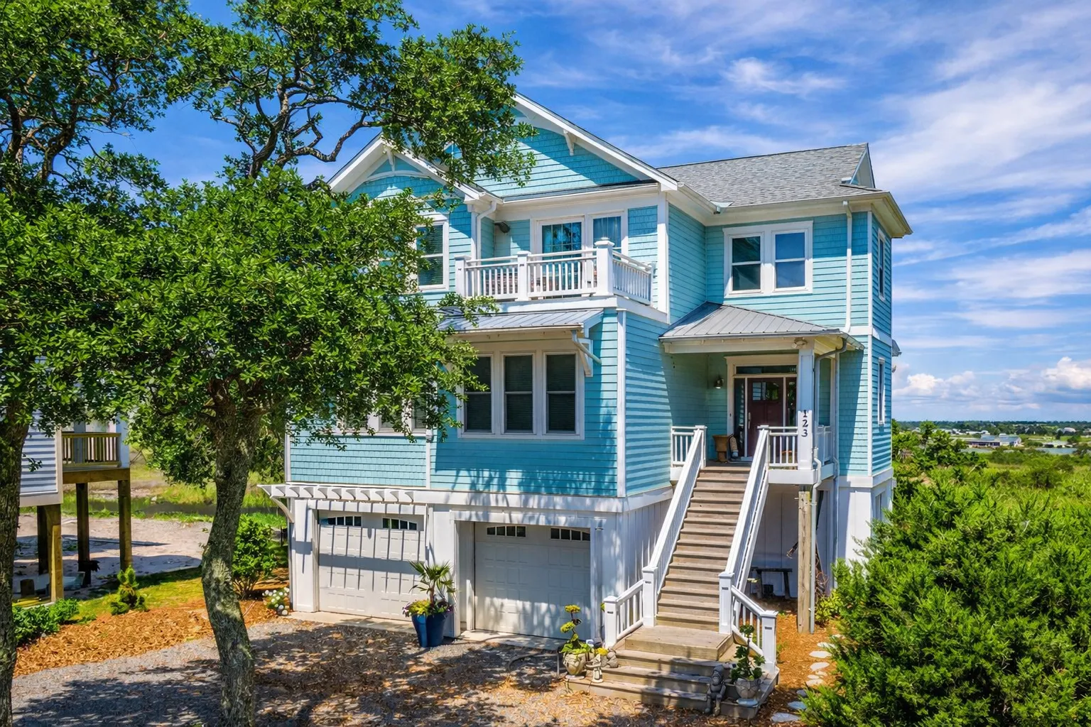 Front exterior view of an elevated coastal beach house with blue lap siding, covered porches, exterior stair entry, and garage below designed for flood-prone coastal living
