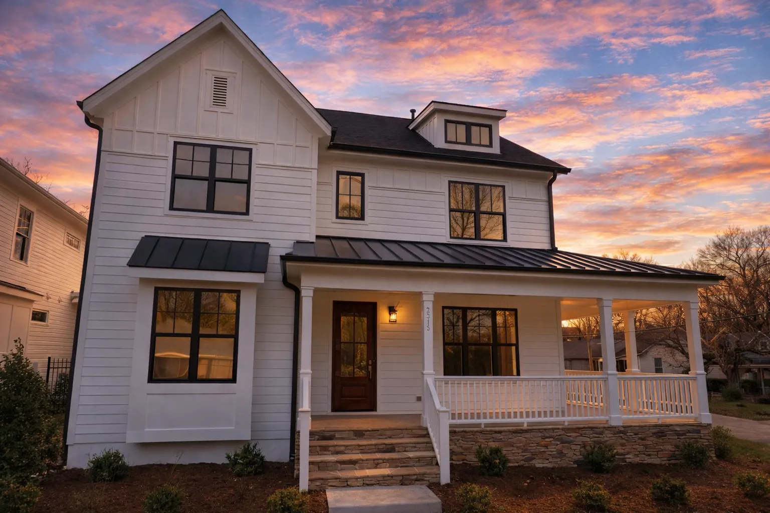 Front elevation of a modern farmhouse with white horizontal lap siding, black windows, covered porch, and standing seam metal roof at sunset
