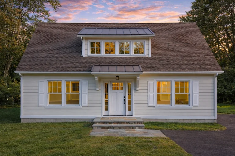 Front elevation of a Cape Cod Cottage featuring navy blue horizontal siding, red-trimmed windows, and a steep roofline surrounded by trees.