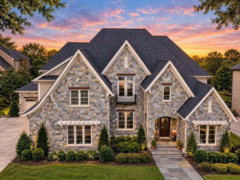 Front exterior of a New American style home with full stone façade, steep gabled rooflines, French Country detailing, and manicured suburban landscaping