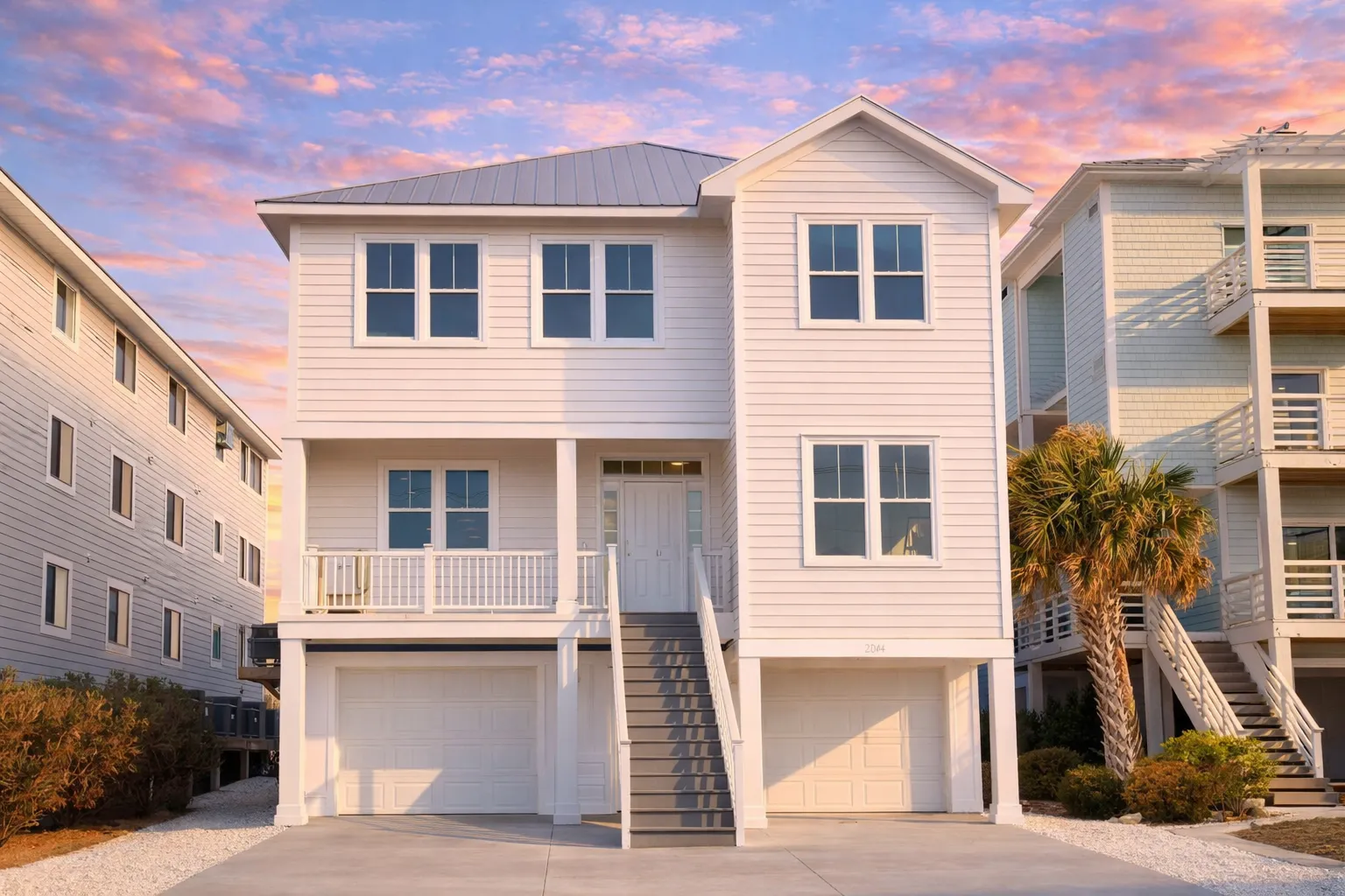 Front elevation of an elevated coastal beach house with horizontal lap siding, standing seam metal roof, exterior stair entry, and garage below