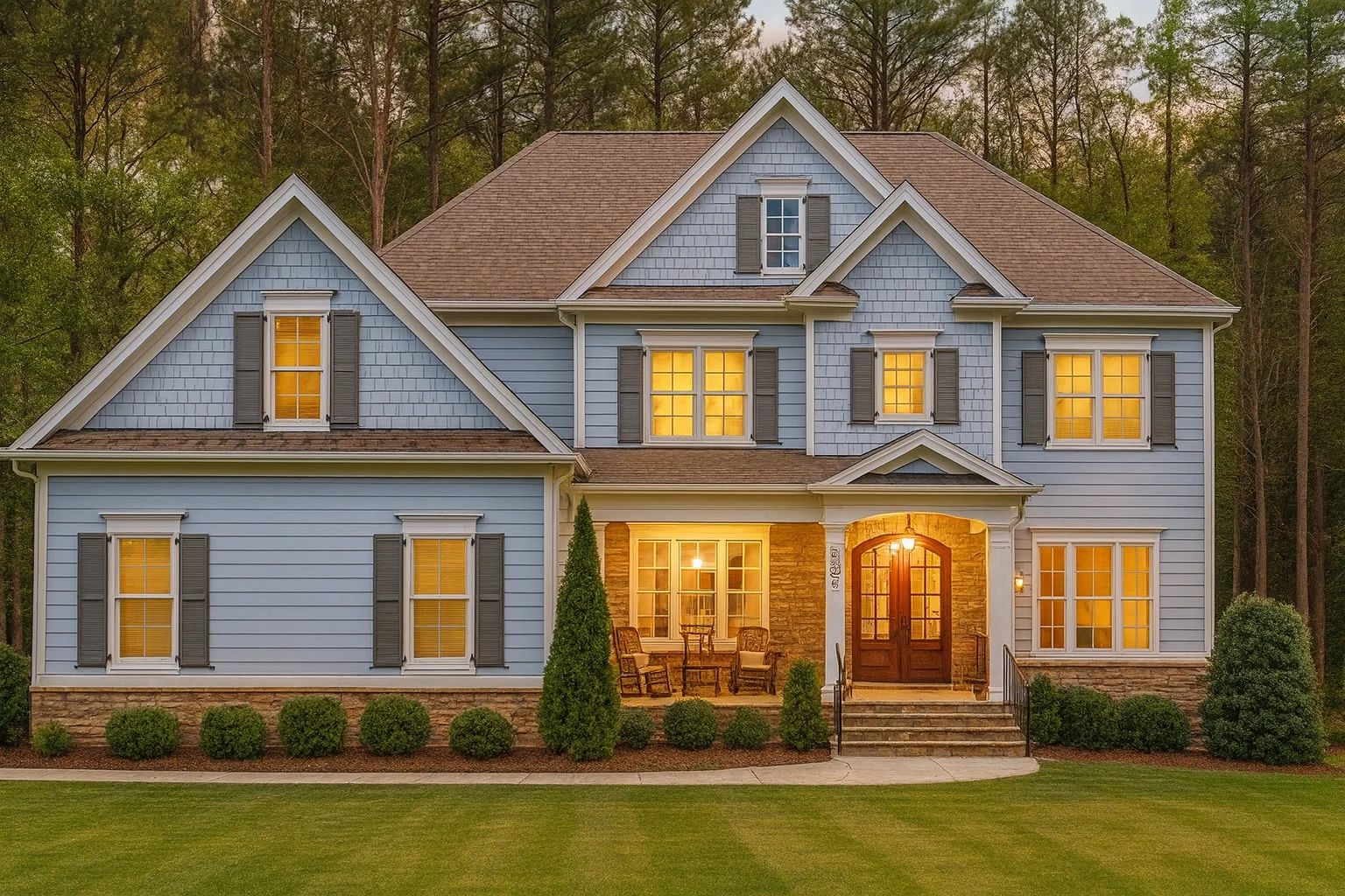 Front exterior of a Shingle Style Coastal home with blue siding, stone porch columns, symmetrical windows, and warm interior lighting